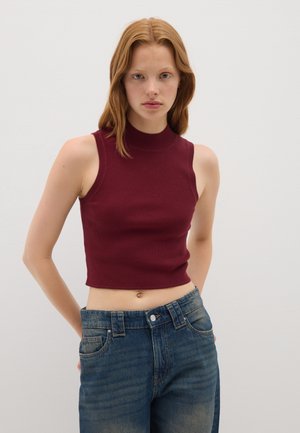 Top corto senza maniche bordeaux con collo alto, texture a coste, abbinato a jeans in denim blu a vita alta con chiusura a bottone e tasche.