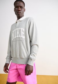 Siv sweatshirt z velikim belim logotipom "NIKE" in sprednjim žepom, v kombinaciji s svetlo roza kratkimi hlačami, na nevtralni podlagi.