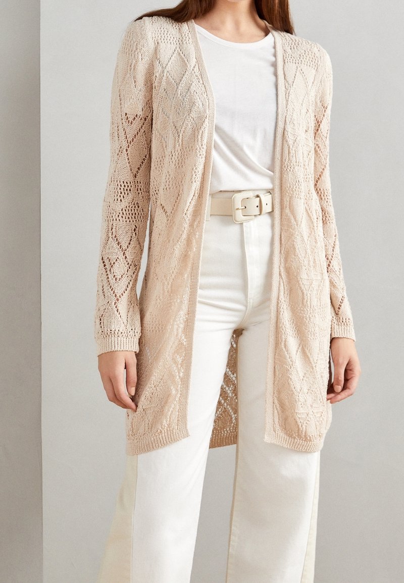 Kobieta w beżowym sweterku ażurowym noszonym na białej koszulce, włożonej do białych spodni z beżowym paskiem, na jednolitym tle.