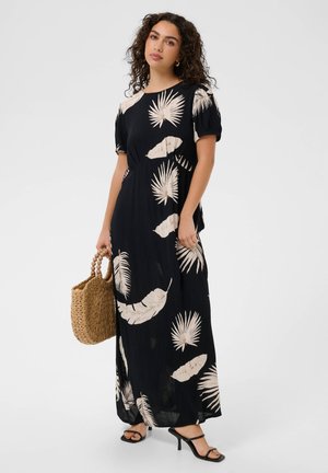 Kobieta ubrana w czarną sukienkę maxi z beżowym tropikalnym wzorem liści, trzyma plecioną słomianą torebkę i nosi czarne sandały na obcasie.
