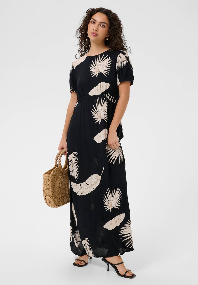 Femme portant une robe longue noire avec des imprimés de feuilles tropicales beiges, tenant un sac en paille tressé et portant des sandales noires à talons.