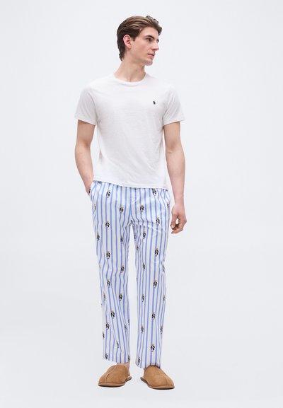 Άνδρας που φοράει λευκό κοντομάνικο μπλουζάκι, μπλε και άσπρη ριγέ πυτζάμα με μικρά πατρόν αρκούδων και καφέ παντόφλες, στέκεται με τα χέρια στις τσέπες.