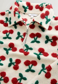 Kremna flis pulover s četrtinskim zadrgo, okrašen z rdečimi cvetličnimi vzorci z zelenimi listi in srebrno zadrgo. Na etiketi piše »NEXT«.