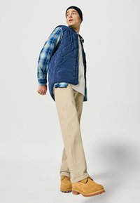 Navy quilted vest over a plaid shirt, beige pants, and yellow-brown boots. The vest features a zip closure and side pockets.
