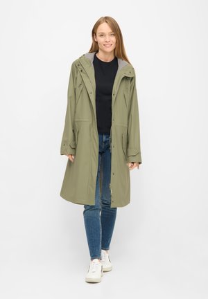 Jeune femme marchant avec un imperméable à capuche vert olive, un chemisier noir, un jean bleu et des baskets blanches sur fond blanc.