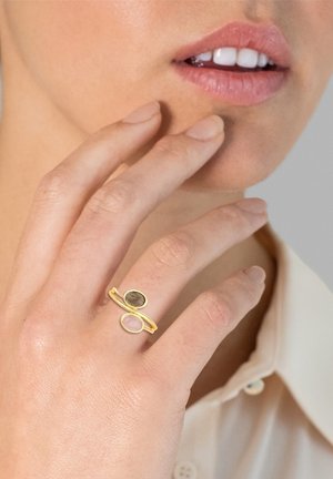 Goldring mit zwei ovalen Steinen, einem grauen und einem rosa, mit einem minimalistischen offenen Design an einem Finger. Glatte Metall- und Edelsteinoberflächen sind sichtbar.