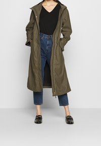 Olivově zelený dlouhý kabát s kapucí, vyrobený z hladkého materiálu, s předním zipem a bočními kapsami, nošený přes tmavé džíny.