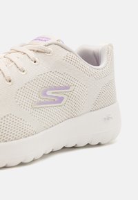 Light beige athletic shoe featuring a breathable mesh upper, textured pattern, purple logo detail, and a cushioned white sole.
