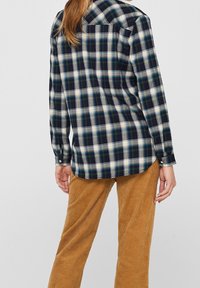 Camicia a quadri in blu scuro, bianco e teal, con maniche lunghe e orlo curvo. Abbinata a pantaloni in corduroy color sabbia.