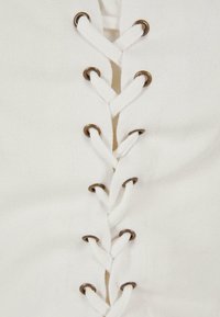 Vitt tyg med en texturerad yta har en snörningsdesign i mitten, hålls av metalliska öglor. Snörningarna är gjorda av tjock vit snodd.