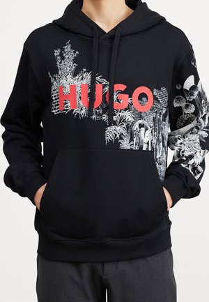 Czarna bluza z kapturem z czerwonym napisem "HUGO" i białą grafiką inspirowaną naturą na piersi i prawym rękawie, noszona przez osobę z rękami w kieszeniach.