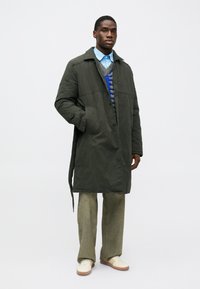 Lång olivgrön kappa med robust textur och framfickor, lager-på-lager över en blå tröja och skjorta med krage, kombinerad med ljusgröna byxor och vita skor.