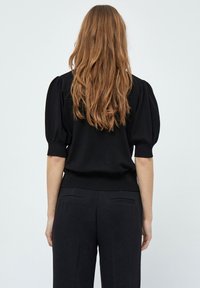 Žena s dlouhými světlými hnědými vlasy stojí zády, oblečená v černém topu s krátkým rukávem a nabíranými rukávy a černých kalhotách na jednobarevném pozadí.