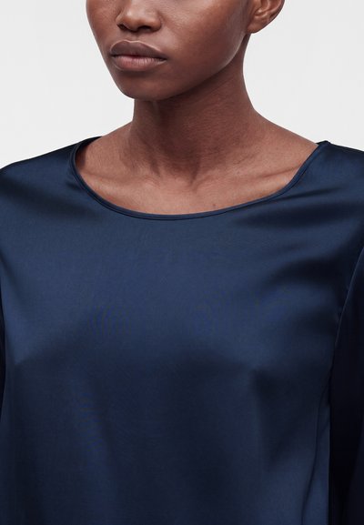 Gros plan d'une personne portant un chemisier en satin bleu foncé avec un large col rond, montrant le cou et le haut de la poitrine.