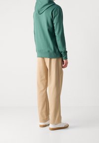Felpa verde in cotone con cappuccio, abbinata a pantaloni beige a gamba larga e sneakers bianche con suola color nocciola. Il tessuto appare morbido e informale.