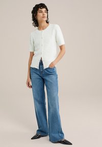 Kortärmad, elfenbensfärgad, fluffig cardigan med rund halsringning, knappstängning och fransade kanter, tillsammans med ljusblå vida jeans och spetsiga svarta skor.