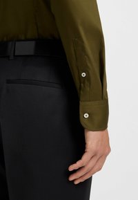 Olijfgroene blouse met lange mouwen en een stijlvolle, gladde textuur. Voorzien van een zwarte manchetknop en een op maat gemaakte zwarte broek met een achterzak.
