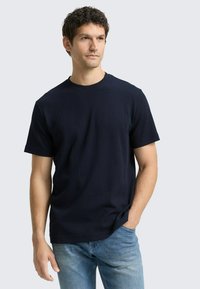 Granatowy t-shirt z krótkim rękawem, okrągłym dekoltem i prążkowaną fakturą. Noszony z jasnoniebieskimi dżinsami, prezentującymi swobodny krój.