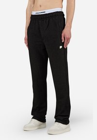 Pantalons de survêtement noirs côtelés avec une ceinture élastique arborant un logo blanc au niveau de la hanche. Portés avec des sneakers blanches, mettant en avant une coupe décontractée.