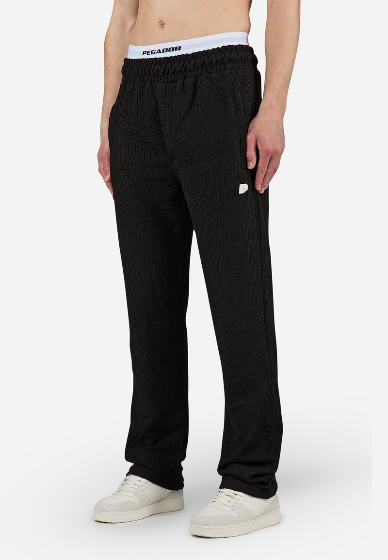 Pantalons de survêtement noirs côtelés avec une ceinture élastique arborant un logo blanc au niveau de la hanche. Portés avec des sneakers blanches, mettant en avant une coupe décontractée.