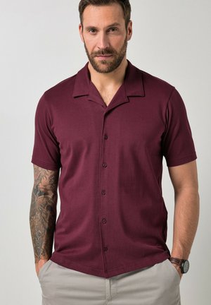 Baarddragende man met getatoeëerde linkerarm draagt een kortarmig bordeaux knoopoverhemd en beige broek, handen in de zakken, neutrale lichte achtergrond.