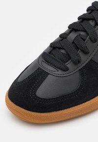 Detailní záběr na černé tenisky s texturovanou kůží a semišovými panely, silnými černými tkaničkami a hnědou gumovou podrážkou.