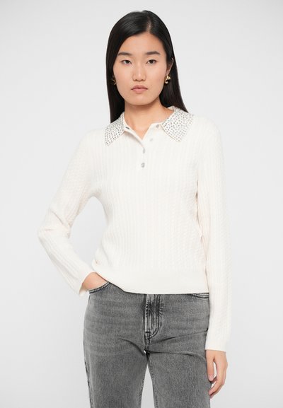 Kvinde iført en hvid kabelstrik sweater med en nitterkrave og grå jeans, stående med den ene hånd i lommen foran en ensfarvet baggrund.