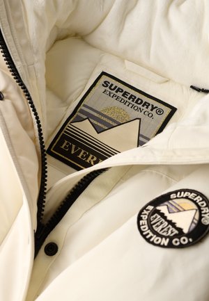 Wnętrze kremowej kurtki z teksturowaną metką z czarnym i złotym wzorem gór, oraz okrągłą naszywką z logo Superdry.