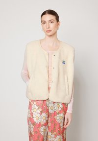 Crème-kleurige fleece vest met een ronde halslijn, drukknoopjes en zijzakken; heeft een klein blauw en groen logo patch.