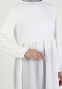 Bílé šaty s dlouhým rukávem a nařaseným pasem, mají hladkou texturu, kulatý výstřih a lehce splývavý střih. Doplněním je světle šedý šátek.