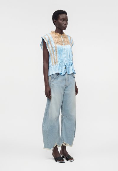 Γυναίκα που φοράει μπλε και μπεζ αμάνικο τοπ με βολάν και κουμπιά, πλατύ παντελόνι τζιν σε ανοιχτό χρώμα με ξεφτισμένες λεπτομέρειες και μαύρα ανοιχτά πέδιλα με τακούνι, όρθια πλάγια.