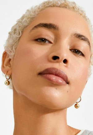 Zelta stīpas auskari ar karājošu sfērisku akcentu. Virsma ir gluda, un kopējais dizains ir minimālistisks, kas papildina seju.