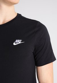 Camiseta de algodón negra con mangas cortas, escote redondo, que presenta un logo de Nike bordado en blanco en el lado izquierdo del pecho. Textura suave.
