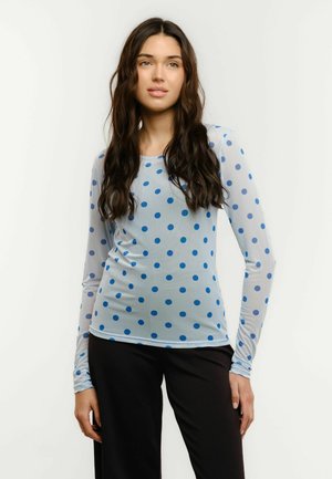 Žena s dlhými tmavými vlnitými vlasmi, oblečená v svetlomodrom tričku s dlhým rukávom s tmavomodrými bodkami a čiernych nohaviciach, stojaca na bielom pozadí.