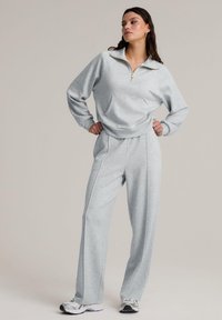 Ljust grå, zip-up sweatshirt med ficka, parat med matchande vida byxor. Materialet verkar mjukt och texturerat, lämpligt för avslappnad användning.