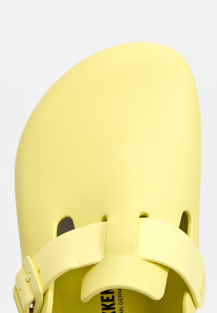 Gelbe Plastiksandale mit abgerundetem Zeh, verstellbarem Riemen und Belüftungsöffnungen. Glatte Textur mit leichtem Design.