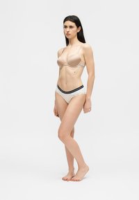 Kvinne med langt mørkt hår som står barfot, iført en nakenfarget BH og hvite truser med en svart elastisk midje.