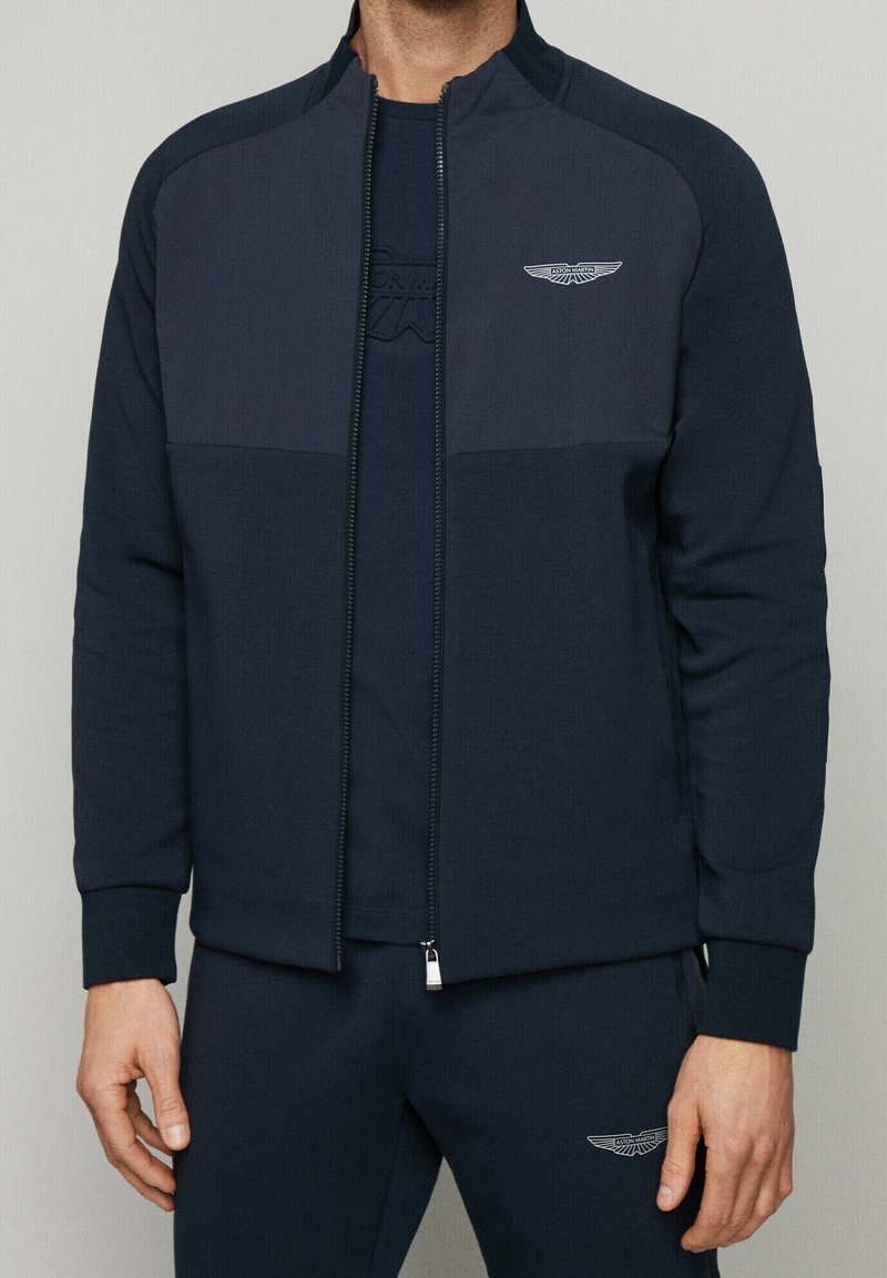 Homme portant une veste zippée Aston Martin bleu marine et un pantalon assorti avec des logos sur la poitrine et la cuisse, debout devant un fond uni.
