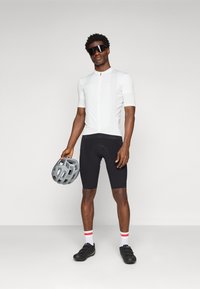 Maillot de cyclisme blanc à manches courtes, short rembourré noir et chaussures de cyclisme noires ; tenant un casque gris ; chaussettes rouges et blanches rayées.