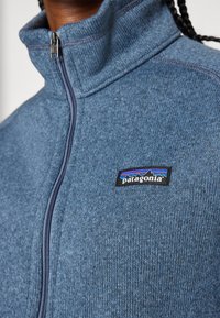 Sinine fliistek jope kõrge kaelusega, millel on täispikk tõmblukk ja must Patagonia logo patch esiküljel. Pehme tekstuur ja helesinine viimistlus.