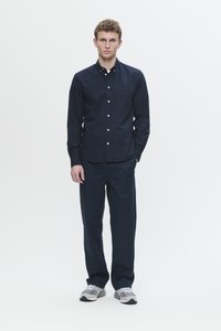 Young man standing facing forward wearing navy button-up shirt, matching navy pants, and gray sneakers against plain white background.