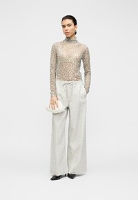 Διαφανές, μπεζ τοπ με λεοπάρ μοτίβο και μακριά μανίκια με ψηλό γιακά· ανοιχτό γκρι παντελόνι με φαρδιά μπατζάκια· κρατάει λευκή υφασμάτινη τσάντα clutch· μαύρα μυτερά παπούτσια.