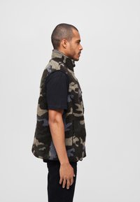 Fleece camouflageväst med hög krage, i nyanser av grönt, grått och blått. Dragkedjefickor på sidorna, strukturerad tyg. Svart t-shirt under.