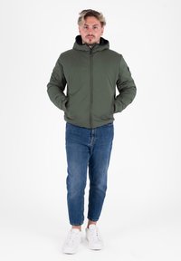 Doudoune vert olive avec fermeture éclair, capuche et poches latérales, portée sur un jean bleu et des baskets blanches sur un fond blanc uni.