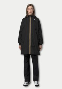 Veste imperméable noire avec capuche, dotée d'une fermeture éclair à l'avant avec des rayures jaunes et blanches et des poches latérales. Portée par-dessus un pantalon noir.