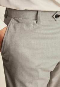 Pantalonii gri deschis au un model herringbone, textură netedă, buzunare laterale, bucle pentru curea și o închidere cu un singur buton la talie.