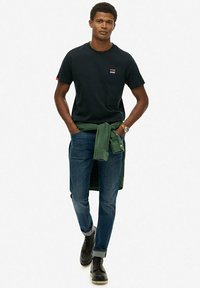 T-shirt nera a girocollo, maglione verde legato in vita, jeans blu e stivali neri con lacci. Outfit semplice e casual dal design pulito.