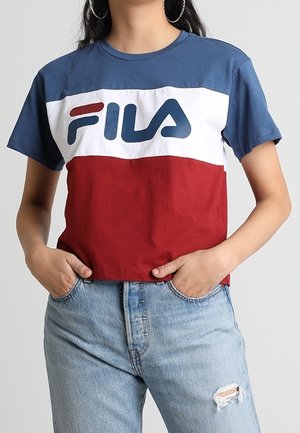 Kobieta w niebiesko-biało-czerwonym krótkim t-shircie Fila, jasnoniebieskich podartych jeansach, z rękami w kieszeniach i kolczykach-kołach.