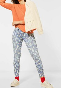 Orange långärmad tröja, krämfärgad jacka, mönstrade blå jeans, röda strumpor och beige skor. Betonar livliga färger och varierande texturer.