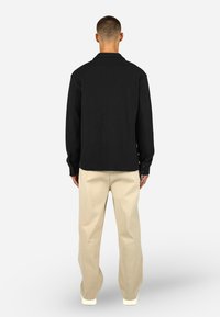 Svart strukturert skjorte med krage, knappede mansjetter, kombinert med beige vide bukser og hvite sneakers; sett fra baksiden.